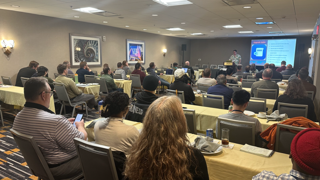A wide shot of a conference presentation room filled with audience members seated at long tables facing front.